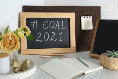 Photo of Chalkboard with hashtag Goal 2021 near tablet and notebook on white table. New year targets