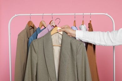 Woman holding wooden hanger with stylish jacket near clothes rack against pink background, closeup