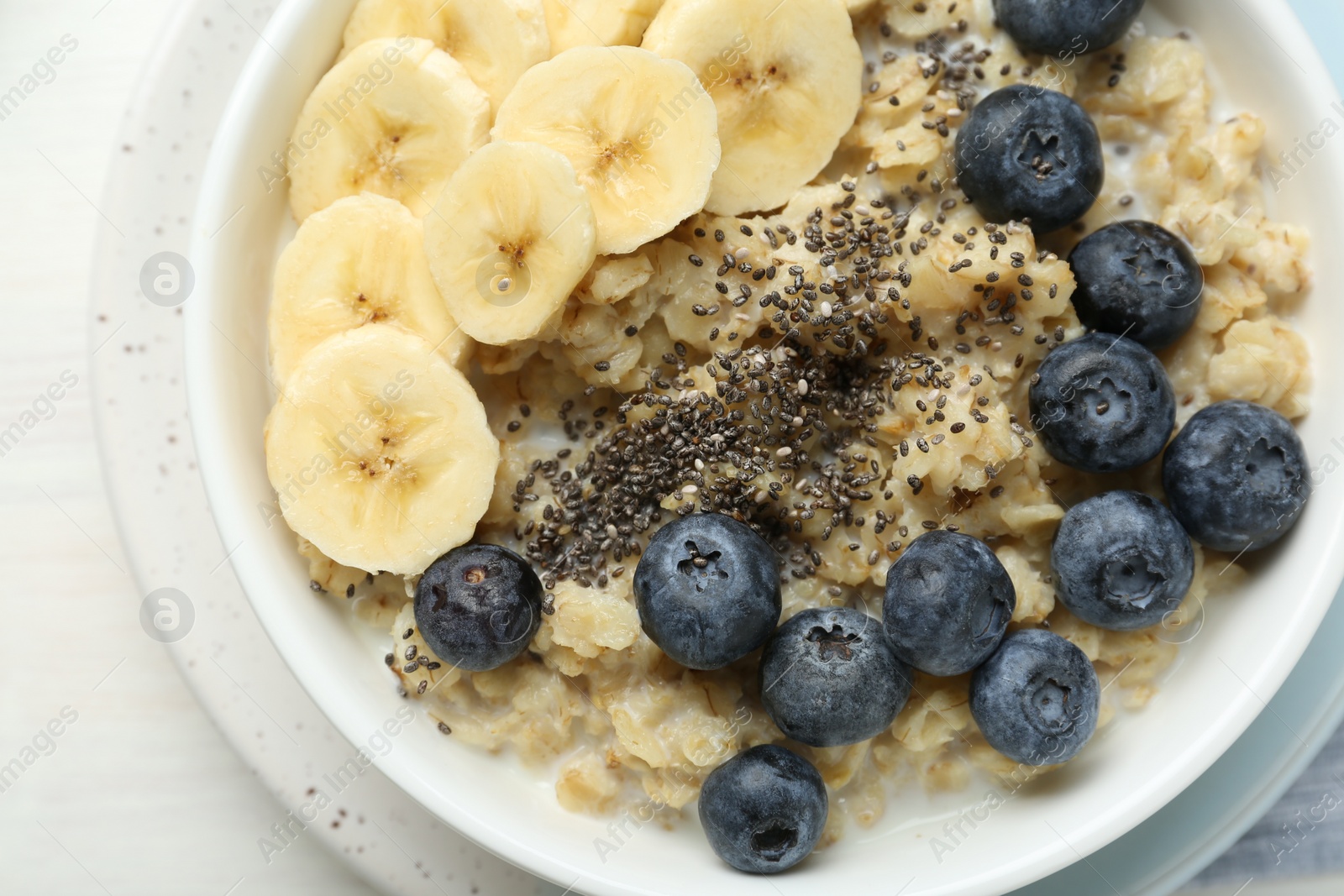 Photo of Tasty oatmeal with banana, blueberries and chia seeds served in bowl on white wooden table, top view