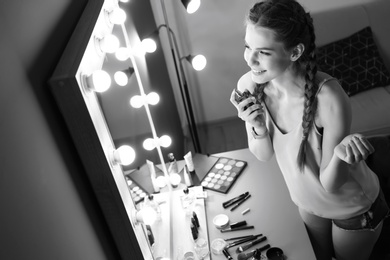 Portrait of beautiful woman with natural makeup applying perfume near mirror indoors, black and white effect