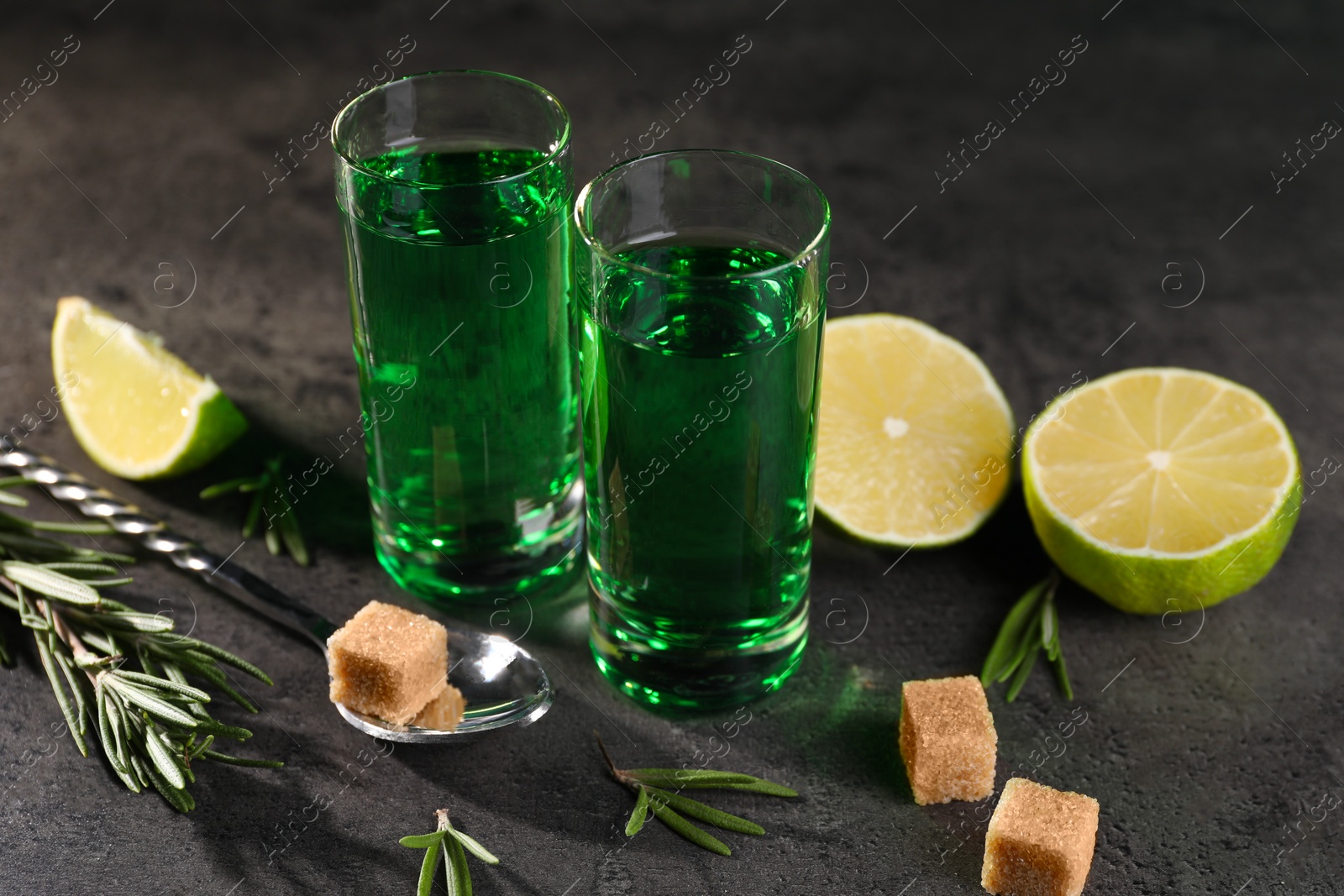 Photo of Absinthe in shot glasses, spoon, brown sugar, lime and rosemary on gray textured table, closeup. Alcoholic drink