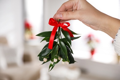 Woman holding mistletoe bunch with red bow indoors, closeup. Traditional Christmas decor