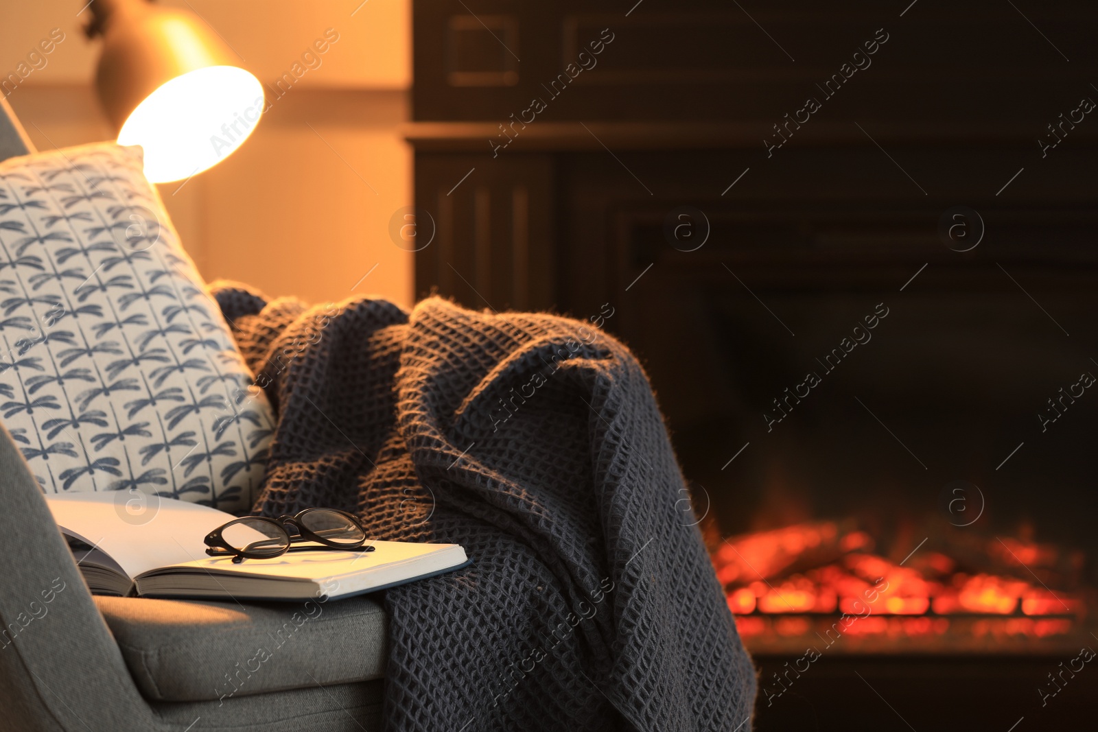 Photo of Open book, glasses and plaid on armchair near fireplace at home, space for text. Cozy atmosphere
