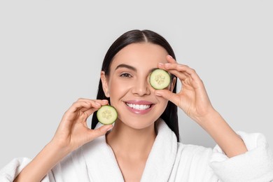 Photo of Woman in bathrobe holding pieces of cucumber on light grey background. Spa treatment