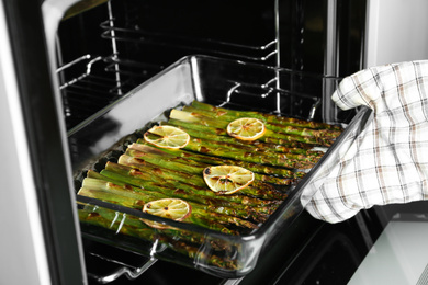 Photo of Person taking glass baking dish with cooked asparagus and lemon slices from oven, closeup