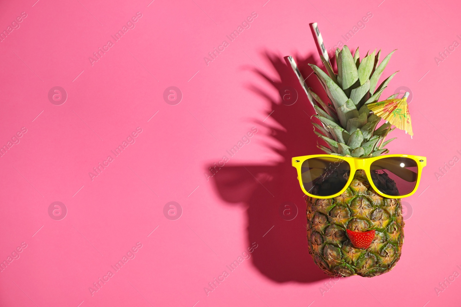 Photo of Funny face made of pineapple, sunglasses and strawberry slice with cocktail umbrella on pink background, top view. Space for text