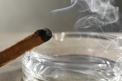 Photo of Smoldering cigar near glass ashtray on light grey table, closeup