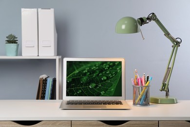 Modern laptop near lamp and holder with stationery on white table