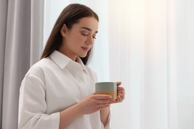 Beautiful woman with cup of drink near window at home, space for text. Lazy morning