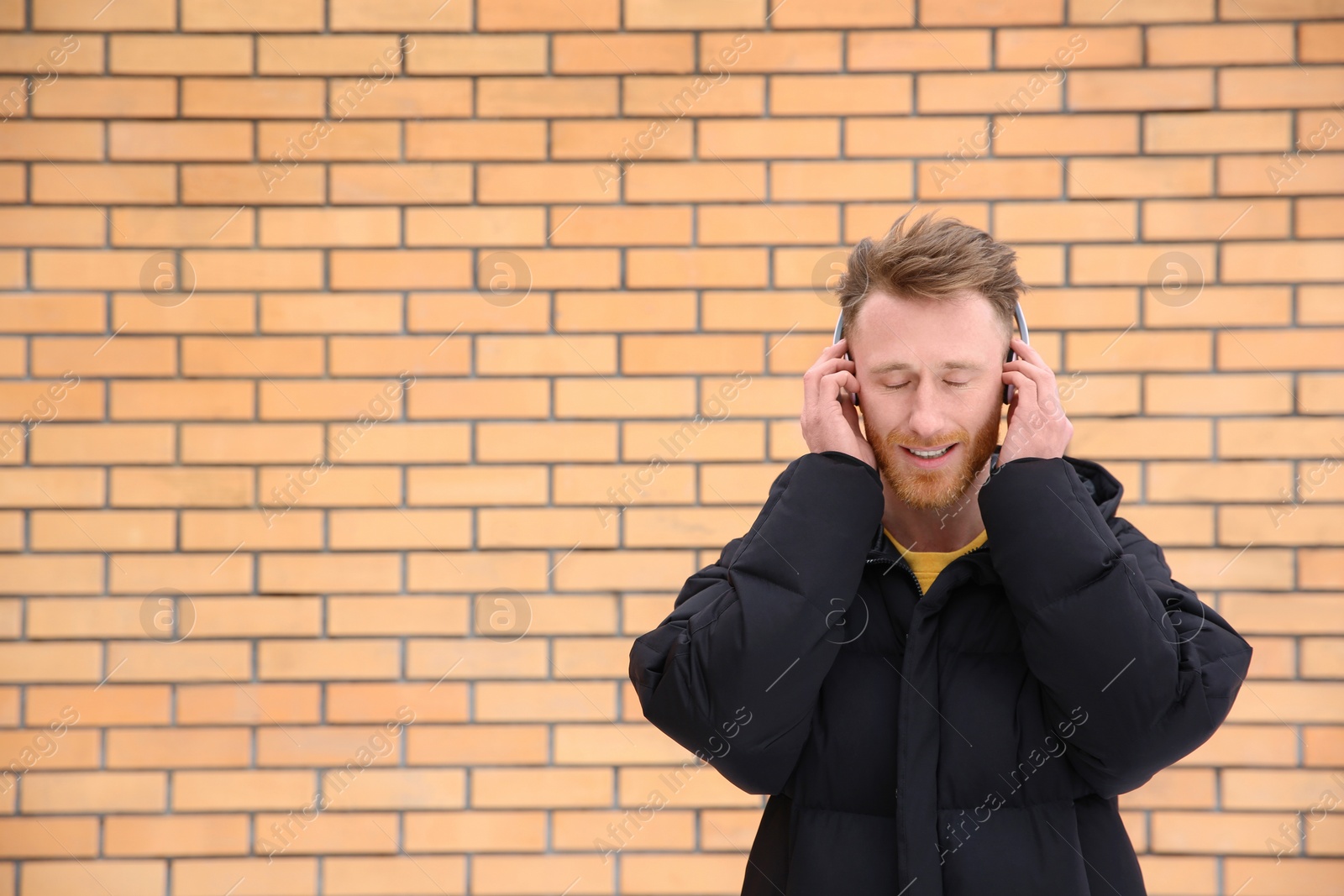 Photo of Young man listening to music with headphones against brick wall. Space for text