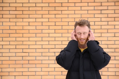 Young man listening to music with headphones against brick wall. Space for text