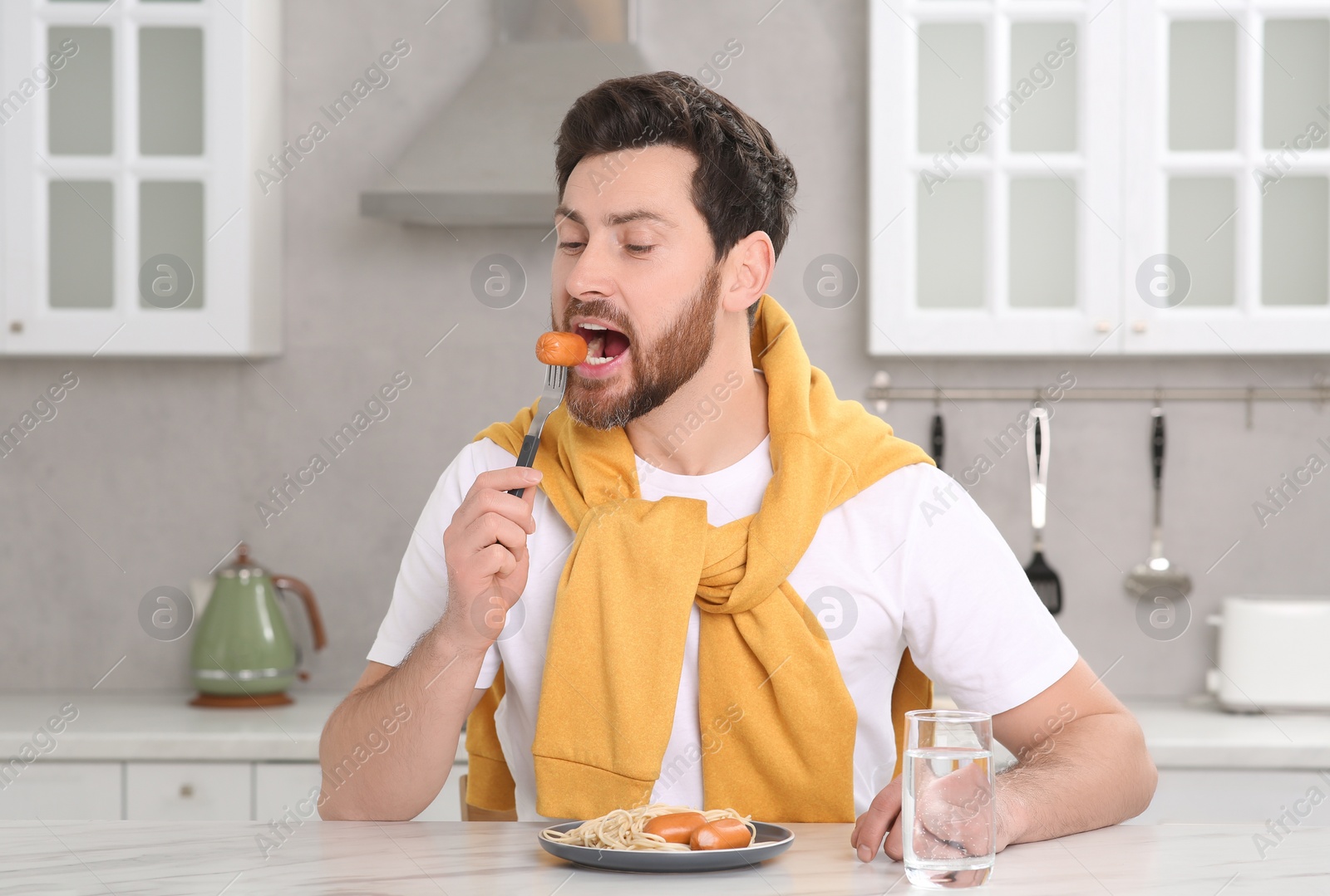 Photo of Man eating sausage and pasta at table in kitchen