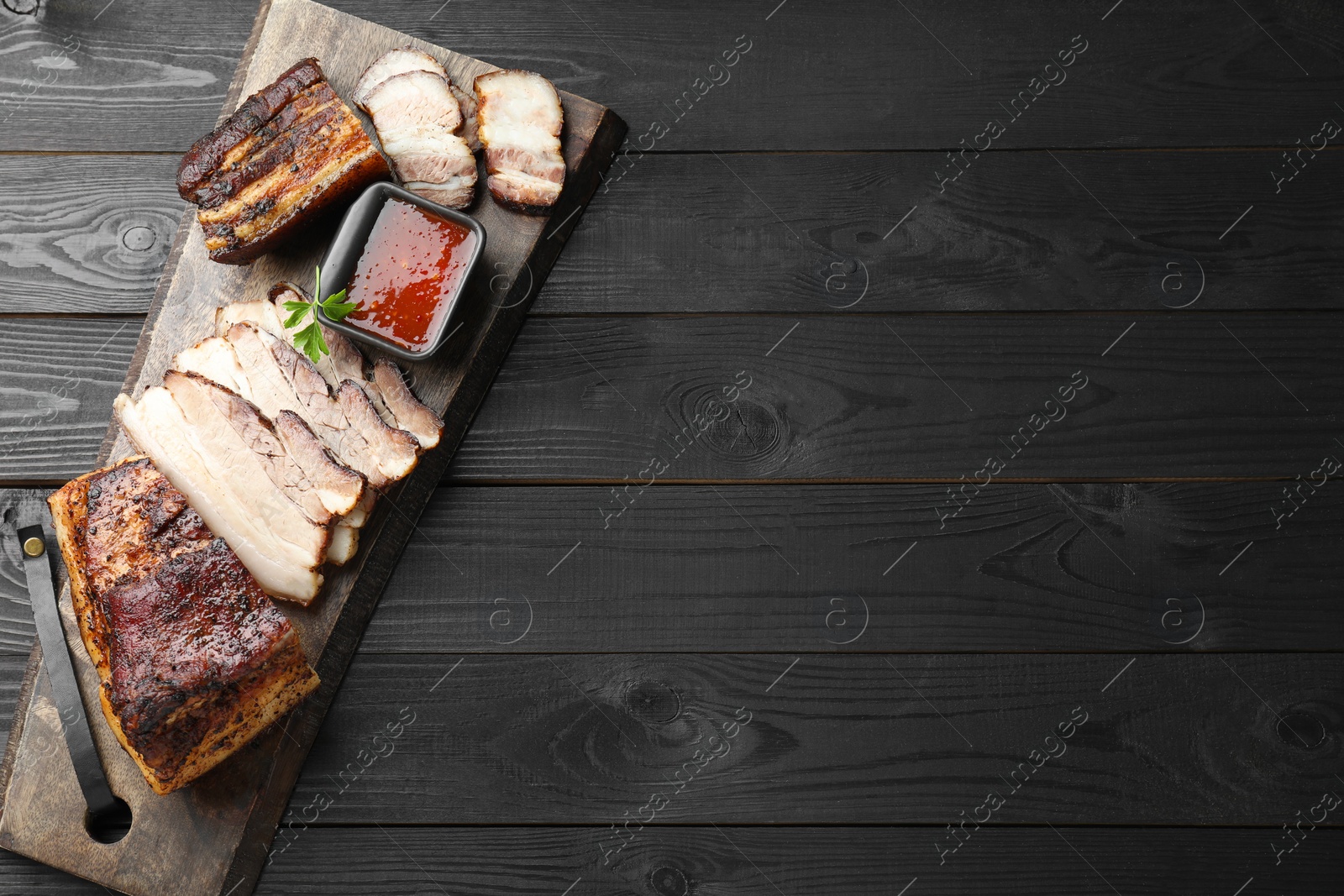 Photo of Pieces of baked pork belly served with sauce and parsley on black wooden table, top view. Space for text