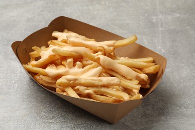 Photo of Tasty potato fries and cheese sauce in paper container on grey table, closeup