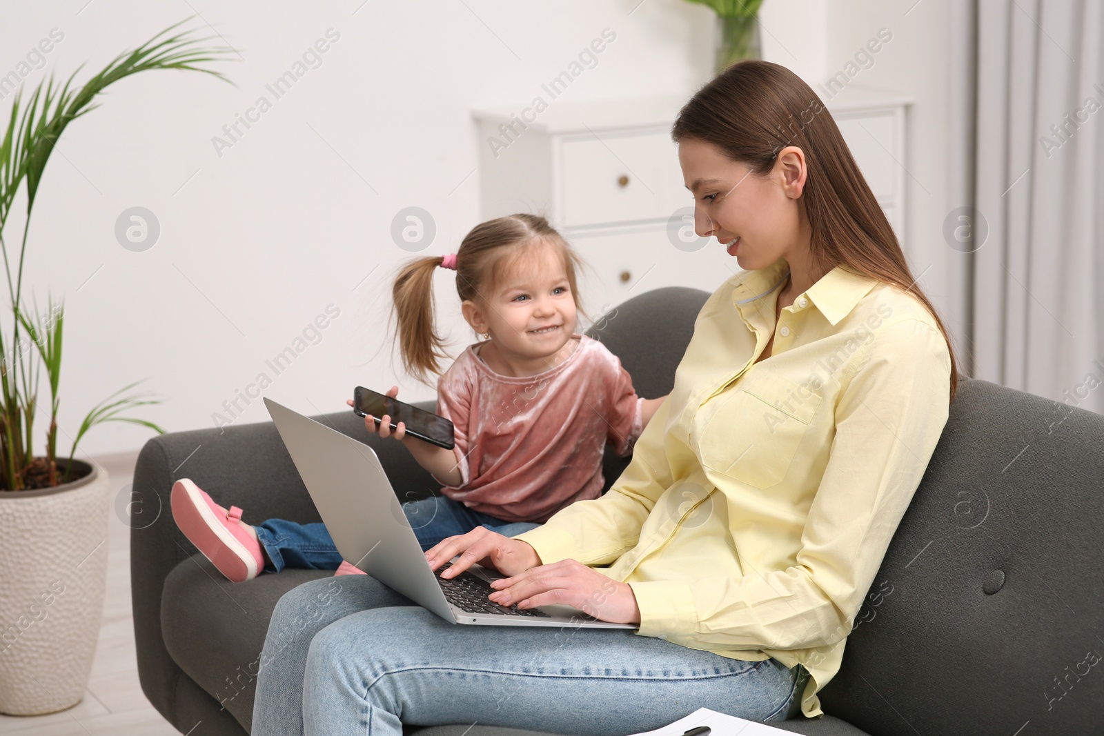 Photo of Mother working remotely on laptop while her daughter playing with smartphone at home