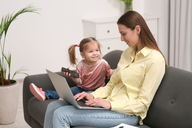 Mother working remotely on laptop while her daughter playing with smartphone at home
