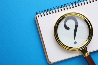 Magnifying glass over notebook with question mark on light blue background, top view. Space for text