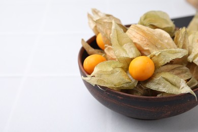 Ripe physalis fruits with calyxes in bowl on white tiled table, closeup. Space for text