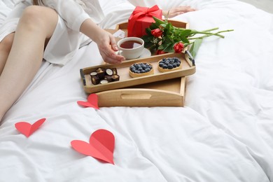 Photo of Tasty breakfast served in bed. Woman with desserts, tea, gift box and flowers at home, closeup