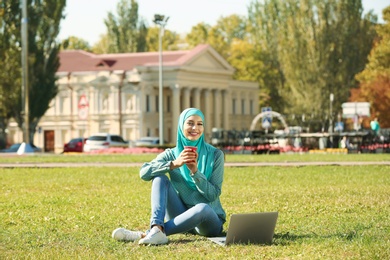 Photo of Muslim woman with cup of coffee and laptop sitting on green lawn outdoors