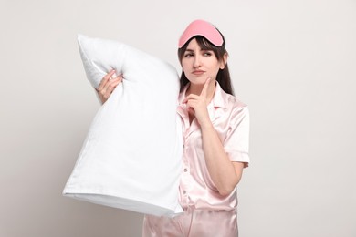 Woman in pyjama and sleep mask holding pillow on light grey background