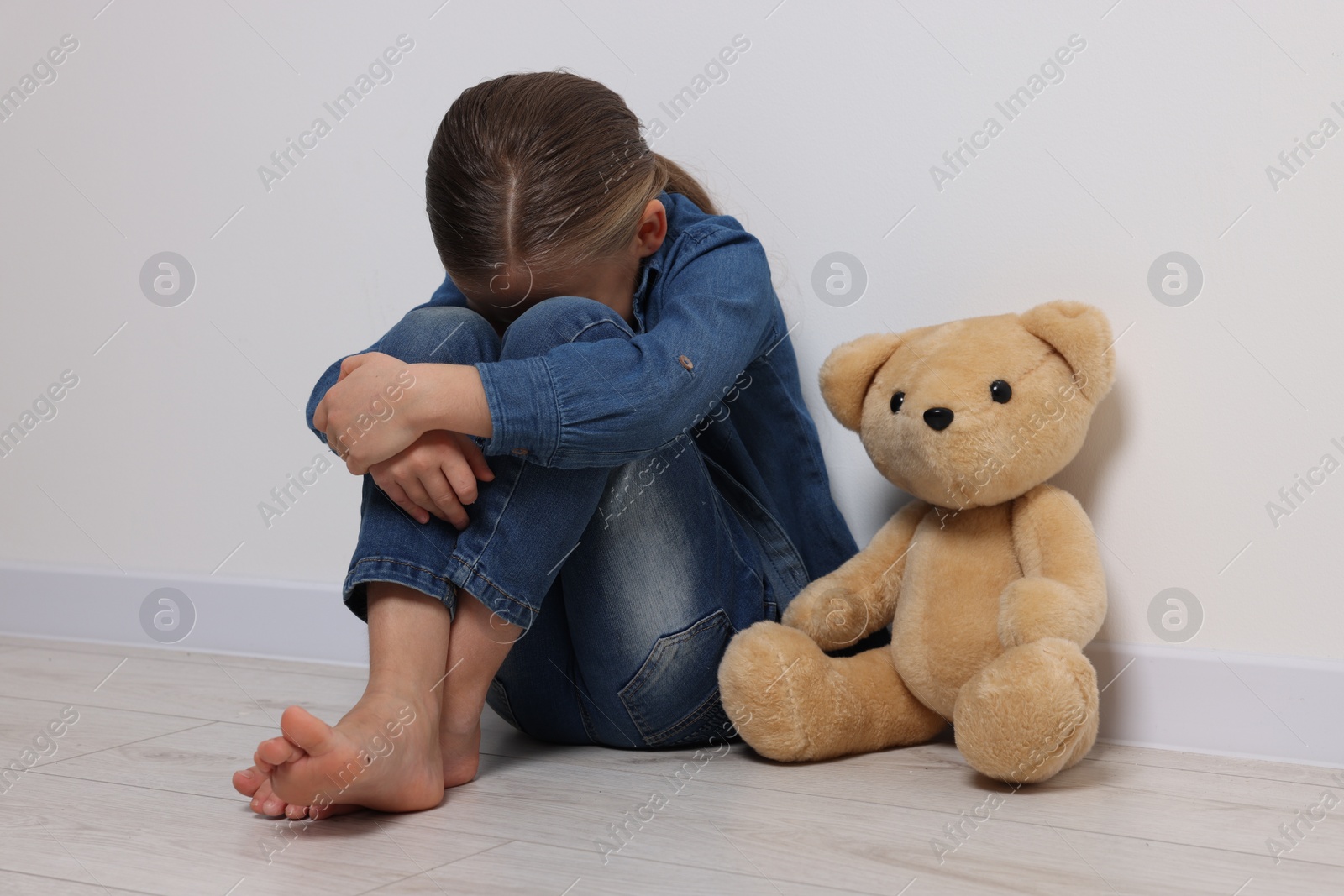 Photo of Child abuse. Upset girl with toy sitting on floor near white wall