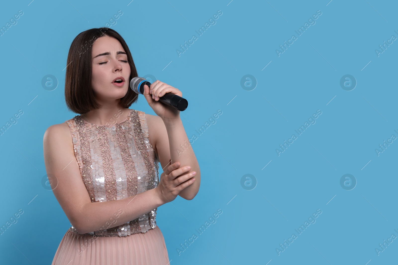 Photo of Beautiful young woman with microphone singing on light blue background, space for text