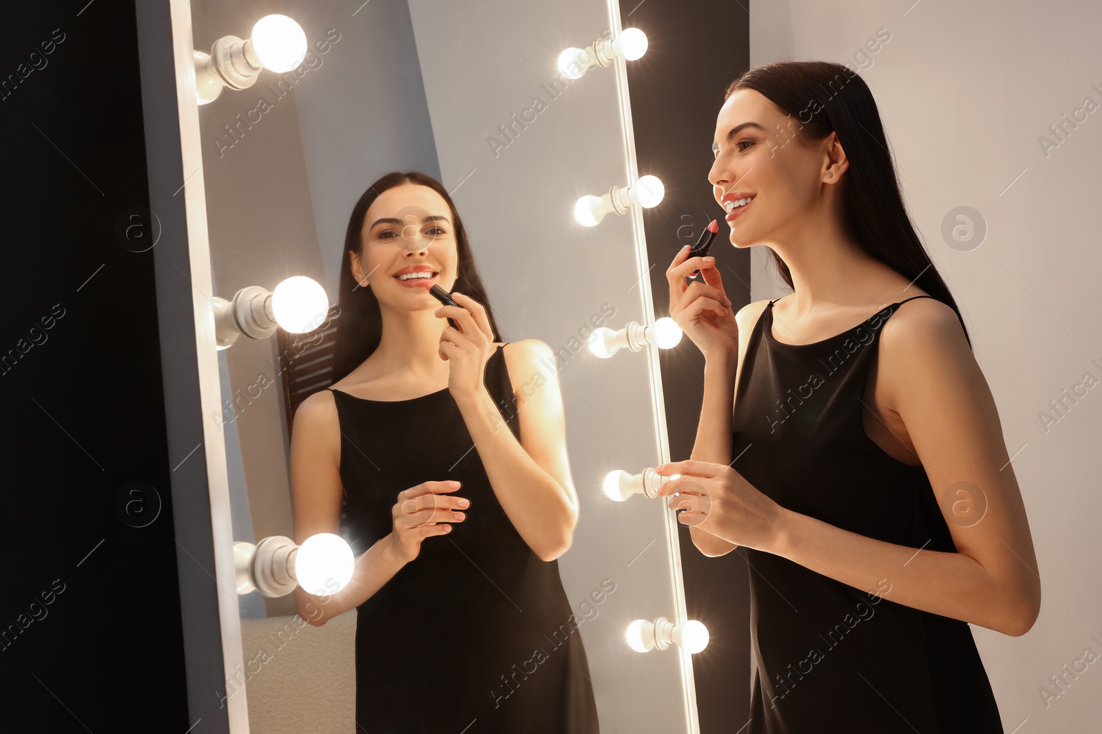 Photo of Beautiful young woman in elegant dress applying lipstick near mirror indoors