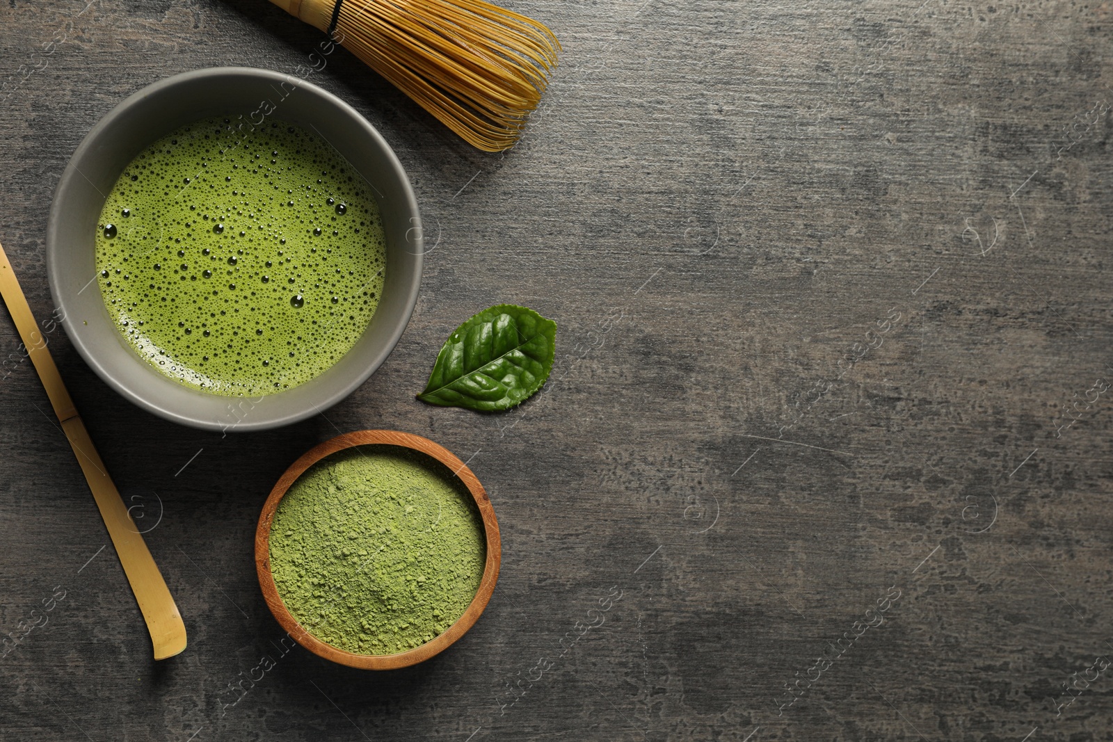 Photo of Cup of fresh matcha tea, bamboo whisk, spoon and green powder on dark grey table, flat lay. Space for text