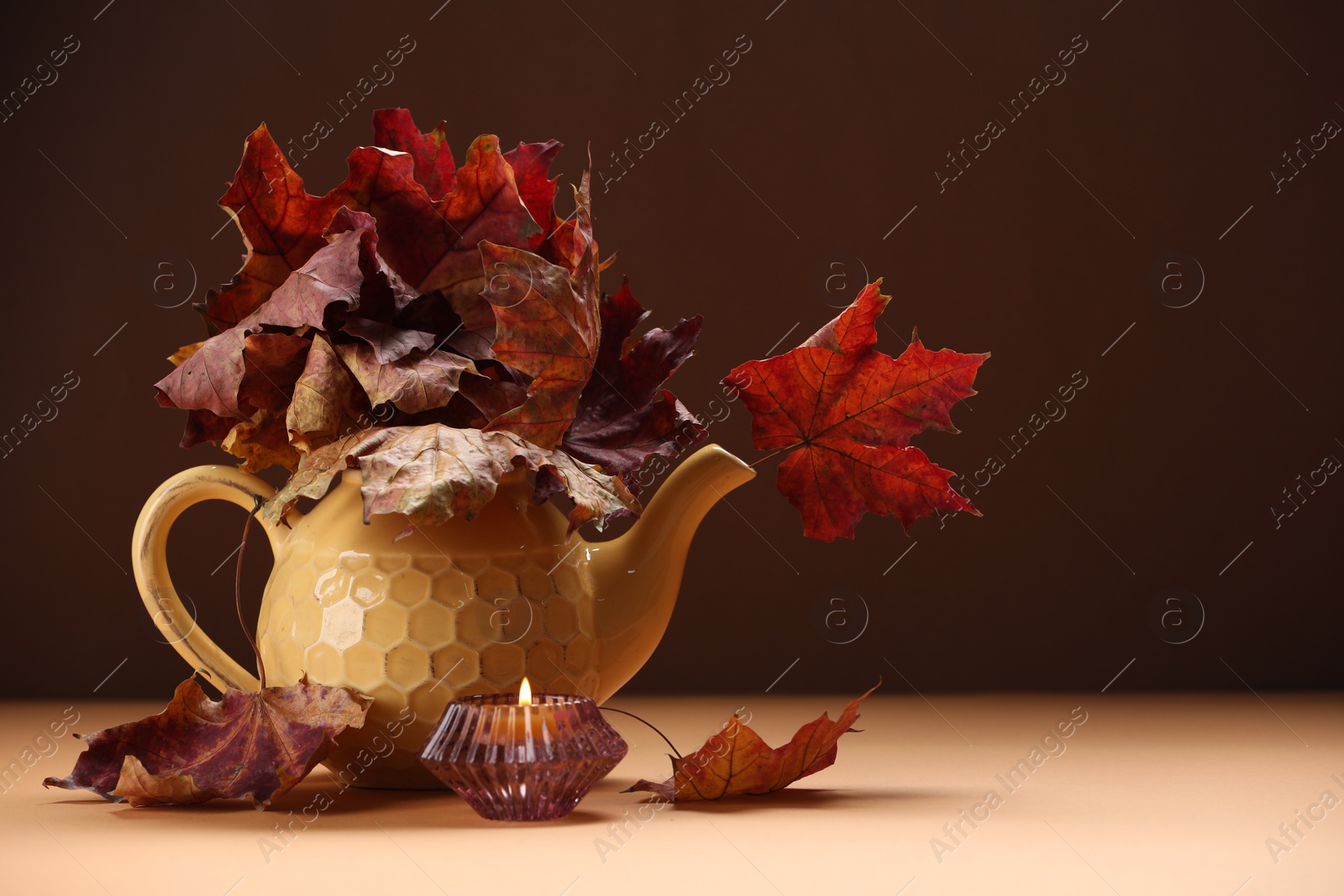 Photo of Teapot with autumn leaves and burning candle on table against brown background