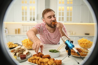 Food blogger eating near microphone at table in kitchen, view through ring light. Mukbang vlog