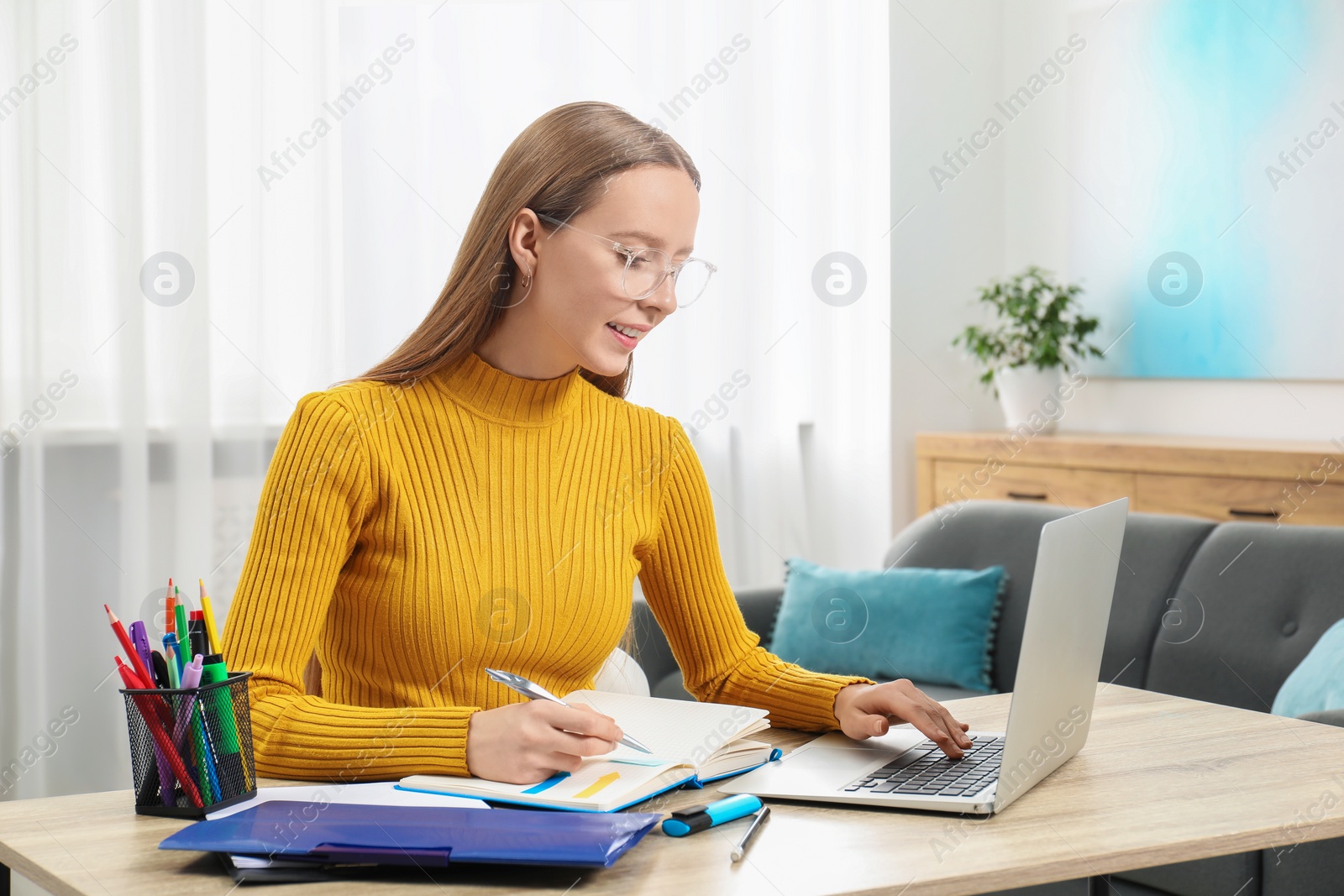 Photo of Woman taking notes while using laptop at wooden table in office