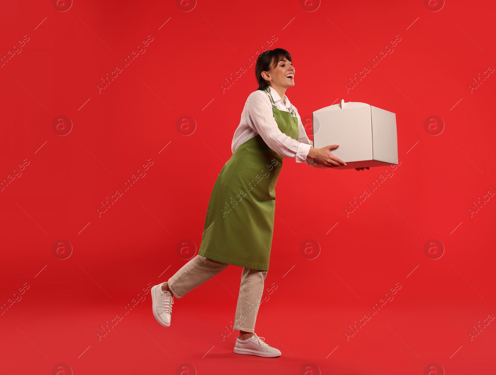 Photo of Happy professional confectioner in apron holding cake box on red background