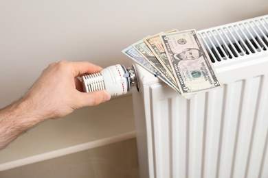 Photo of Man adjusting temperature of heating radiator with money on it indoors