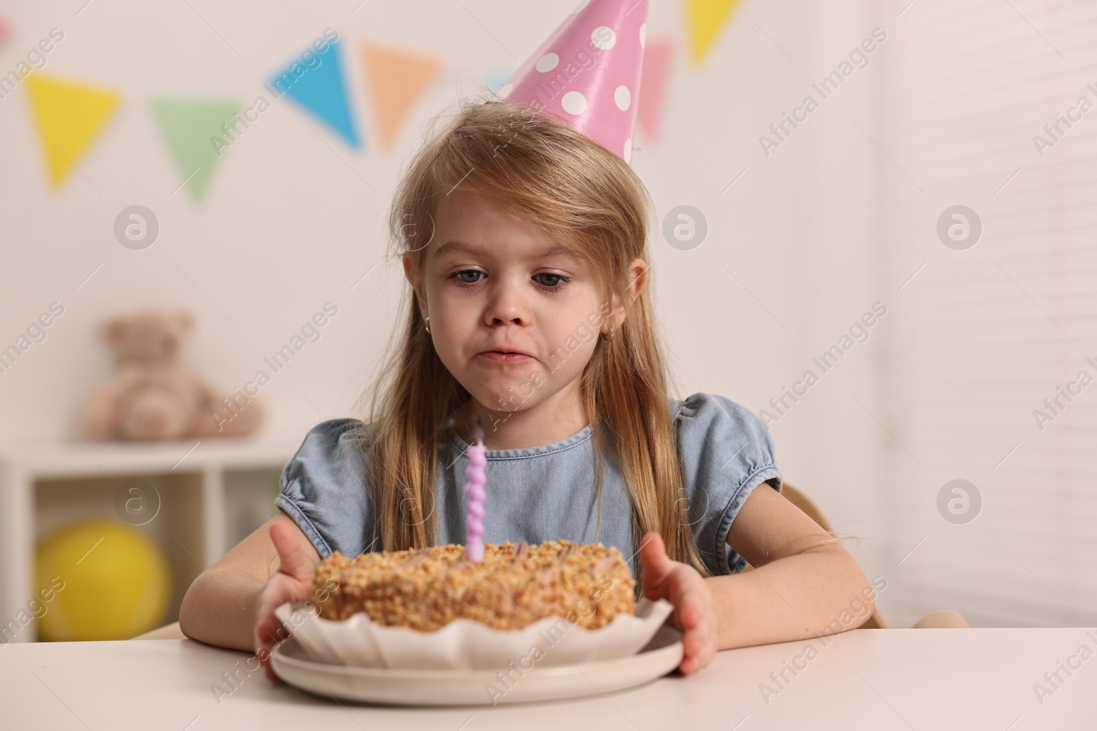 Photo of Cute girl in party hat with birthday cake at table indoors