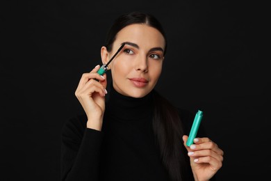 Beautiful young woman applying mascara on black background