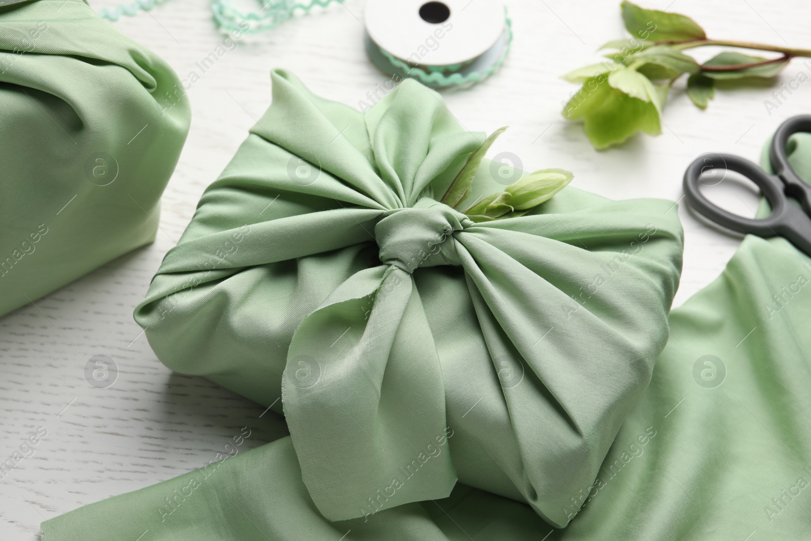 Photo of Furoshiki technique. Gift packed in green fabric and decorated with hellebore flower on white wooden table, closeup