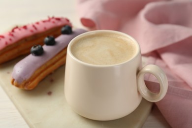 Photo of Cup of coffee and delicious eclairs covered with glaze on white wooden table, closeup