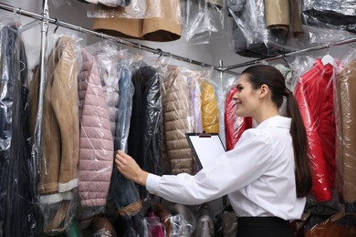 Photo of Dry-cleaning service. Happy worker with clipboard choosing clothes from rack indoors