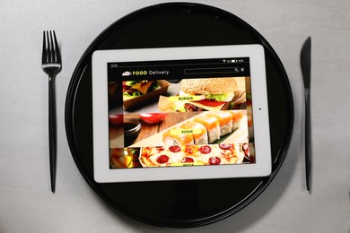 Photo of Modern tablet with open page for online food ordering, plate and cutlery on grey table, flat lay. Concept of delivery service