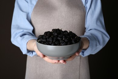 Woman holding bowl of fresh ripe mulberries on black background, closeup