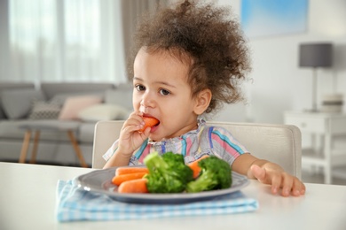 Photo of Cute African-American girl eating vegetables at table in living room
