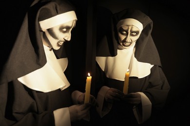 Scary devilish nun with burning candle near mirror on black background. Halloween party look