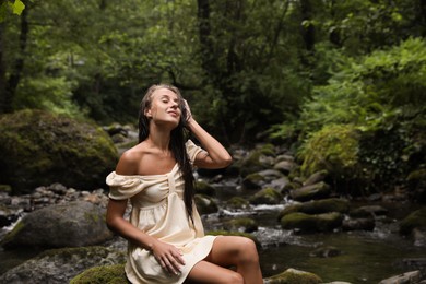 Photo of Beautiful young woman sitting on rock near mountain river in forest. Space for text