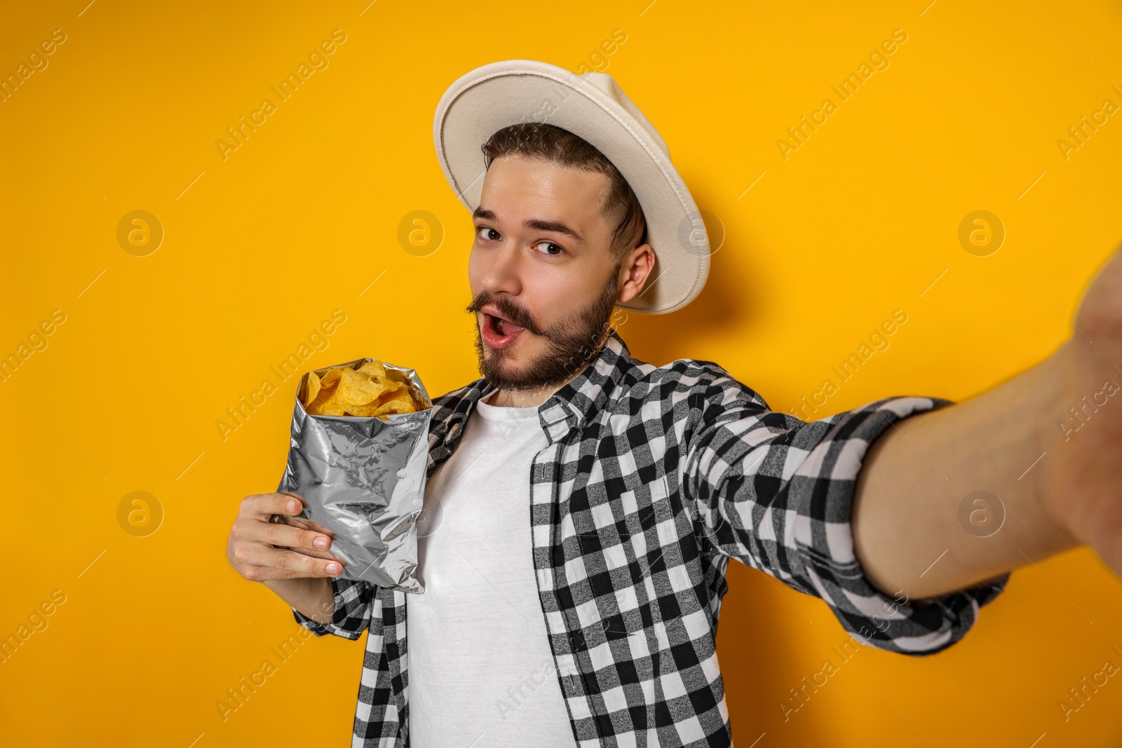 Photo of Handsome young man with bag of tasty potato chips taking selfie on yellow background