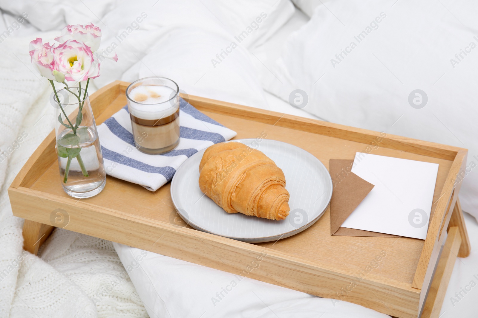 Photo of Tray with tasty croissant, cup of coffee and flowers on white bed