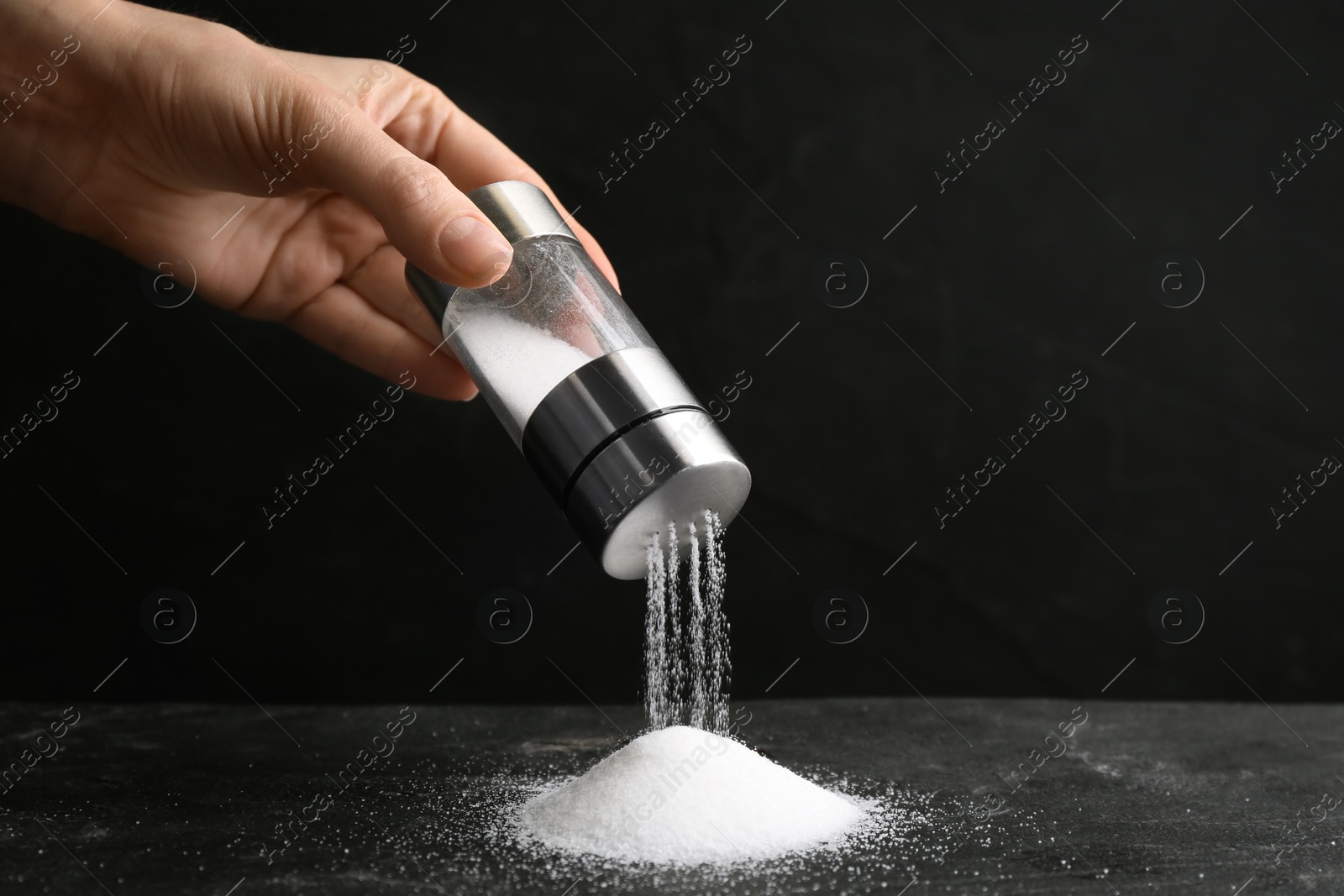 Photo of Woman pouring salt from shaker onto black table, closeup