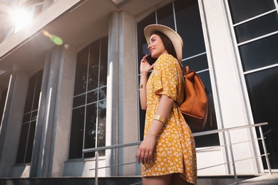 Photo of Beautiful young woman with stylish backpack talking by phone outdoors on sunny day, low angle view