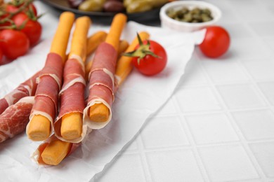 Photo of Delicious grissini sticks with prosciutto and ingredients on white table, closeup. Space for text