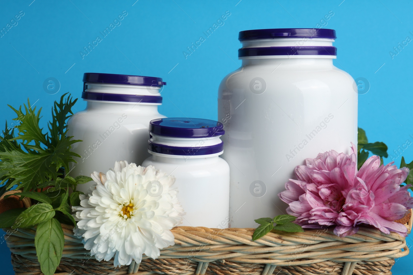 Photo of White medical bottles, arugula and flowers in wicker tray on light blue background, closeup